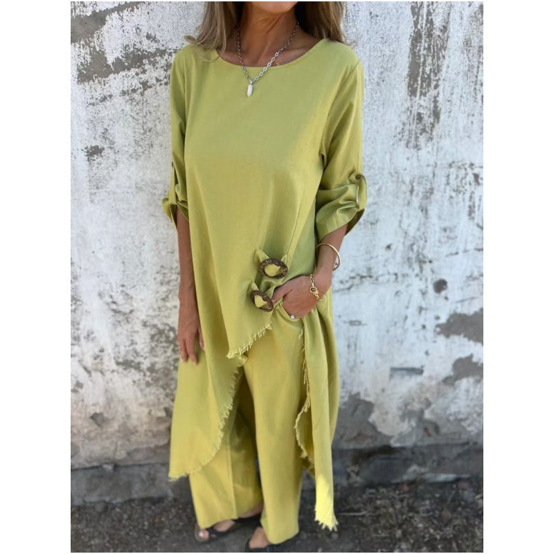 Frau in gelbem, asymmetrischem Leinenkleid mit Fransen, vor einer rustikalen Wand. Modetrend, Sommeroutfit, lässiger Stil.