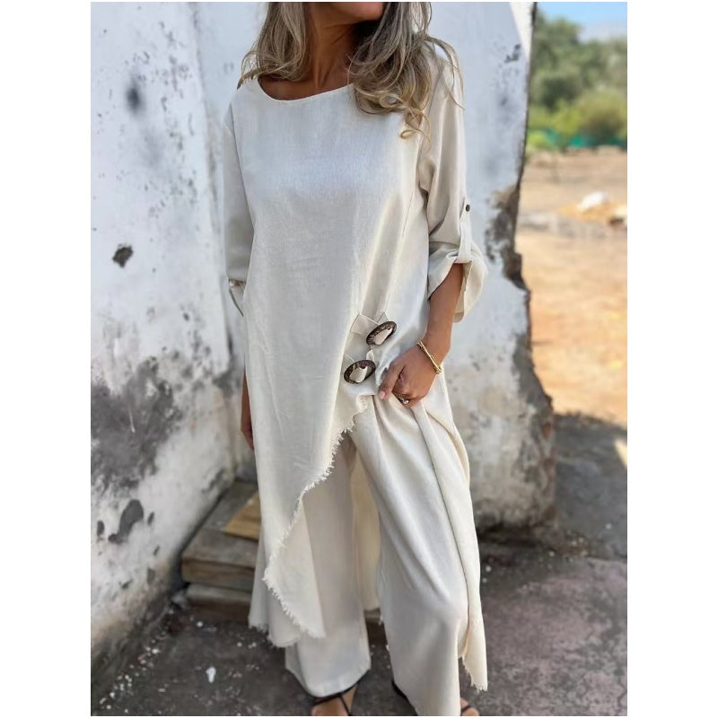 Frau in cremefarbenem, asymmetrischem Leinenkleid mit Knöpfen, steht vor einer verwitterten Wand. Sommermode, lässiger Boho-Stil.
