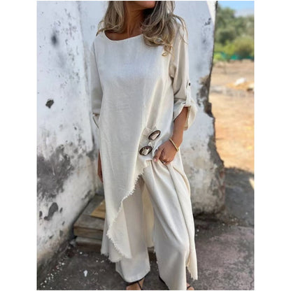 Frau in cremefarbenem, asymmetrischem Leinenkleid mit Knöpfen, steht vor einer verwitterten Wand. Sommermode, lässiger Boho-Stil.