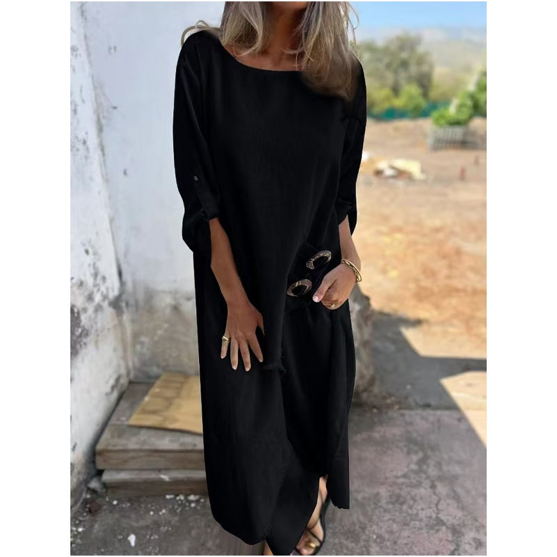 Frau in langem, schwarzem Kleid mit Sonnenbrille in der Hand, steht draußen vor einer weißen Wand. Elegante Sommermode, lässiger Stil.