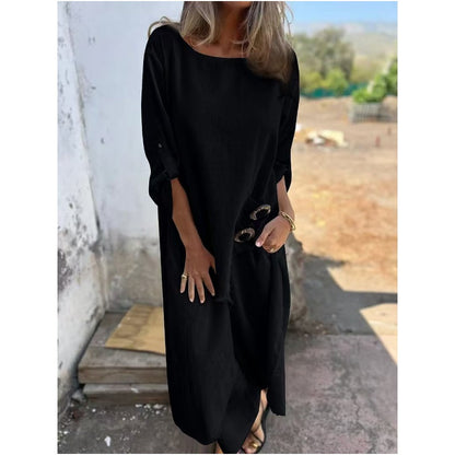 Frau in langem, schwarzem Kleid mit Sonnenbrille in der Hand, steht draußen vor einer weißen Wand. Elegante Sommermode, lässiger Stil.