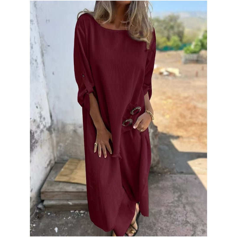 Frau in langem, weinrotem Kleid mit Sonnenbrille in der Hand, steht draußen vor einer weißen Wand. Sommermode, lässiger Stil, elegantes Design.