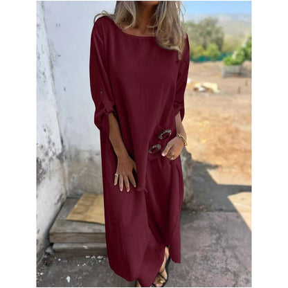 Frau in langem, weinrotem Kleid mit Sonnenbrille in der Hand, steht draußen vor einer weißen Wand. Sommermode, lässiger Stil, elegantes Design.