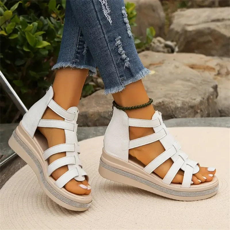 Damenfüße in weißen Sandalen mit Keilabsatz und Riemchen, kombiniert mit zerrissenen Jeans, auf Steinboden im Freien. Modische Sommerschuhe.
