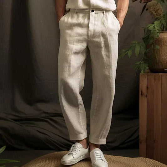 Mann trägt beige Leinenhose und weiße Sneaker, steht vor dunklem Hintergrund mit Pflanzen. Elegante Herrenmode, lässiger Stil, Sommeroutfit.