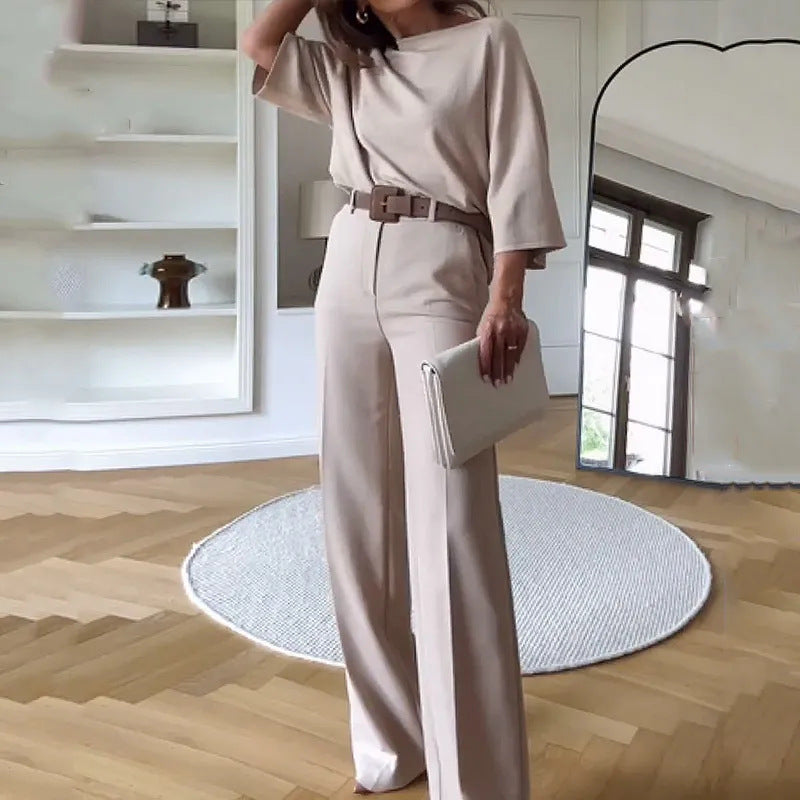 Elegante Frau in beigem Hosenanzug mit Gürtel, hält eine Clutch. Modernes Wohnzimmer mit großem Spiegel und Holzfußboden im Hintergrund.