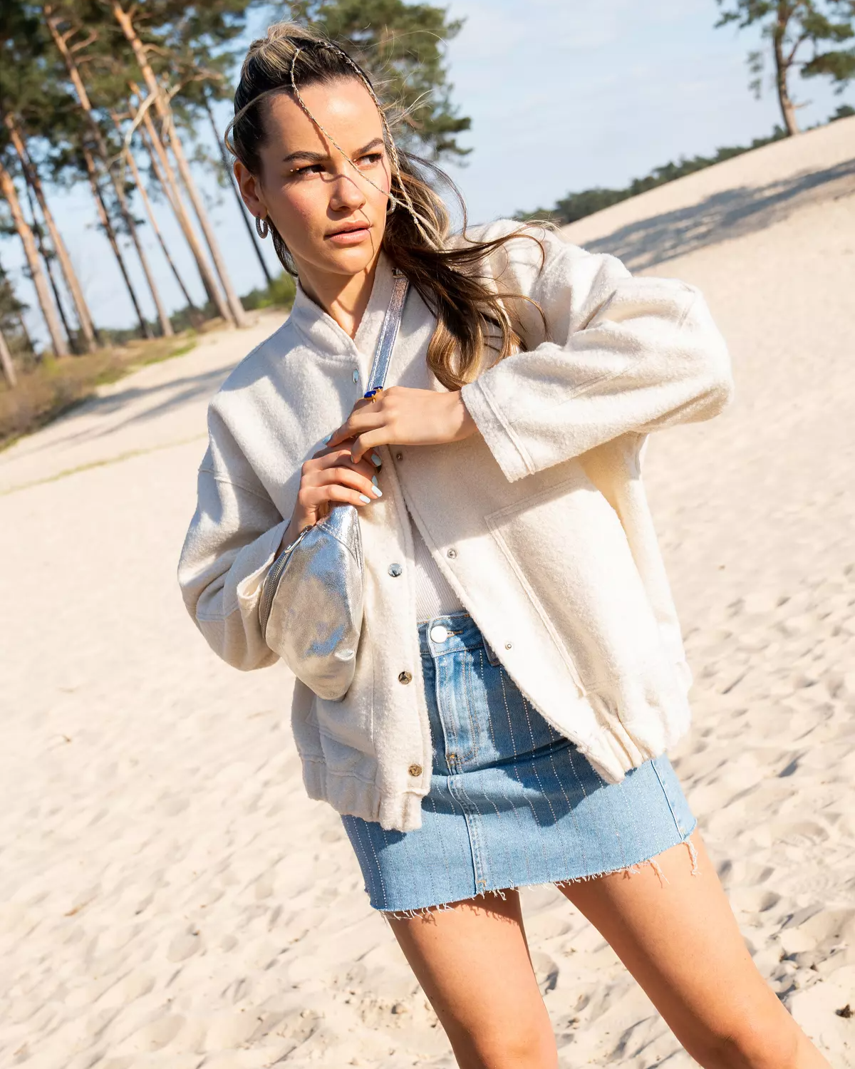 Frau in weißer Jacke und Jeansrock am Sandstrand, mit Bäumen im Hintergrund. Sommermode, Strandspaziergang, Freizeitkleidung.