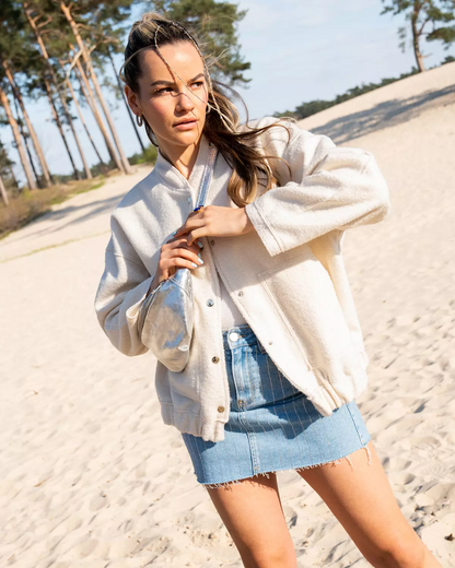Frau in weißer Jacke und Jeansrock am Sandstrand, mit Bäumen im Hintergrund. Sommermode, Strandspaziergang, Freizeitkleidung.