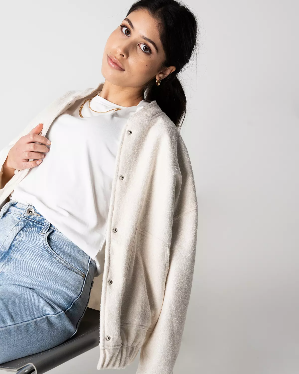 Frau in weißem T-Shirt und Jeans, trägt cremefarbene Strickjacke, sitzt lässig auf Stuhl, modisches Outfit, minimalistischer Stil.