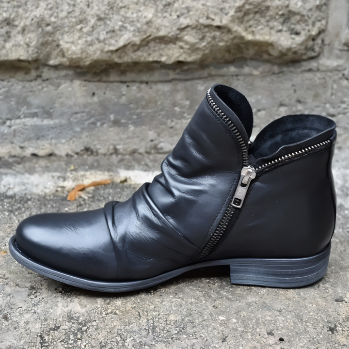 Schwarzer Lederstiefel mit seitlichem Reißverschluss auf grauem Betonboden vor Steinmauer. Eleganter, moderner Schuh für Herbstmode.