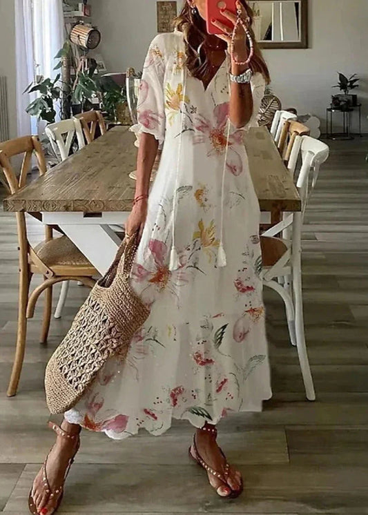 Frau in geblümtem Sommerkleid mit Strohtasche steht in modernem Esszimmer. Holztisch, Stühle, Pflanzen im Hintergrund. Boho-Chic Mode.