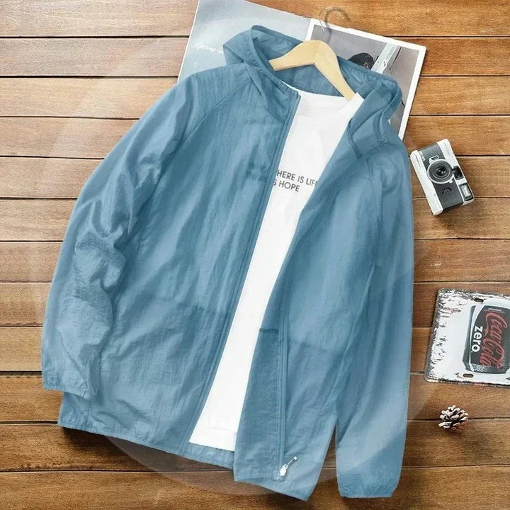 Hellblaue Windjacke mit Kapuze auf Holztisch, daneben weiße T-Shirts, Vintage-Kamera und Coca-Cola-Deckel. Outdoor-Mode, Herrenbekleidung, Freizeitlook.