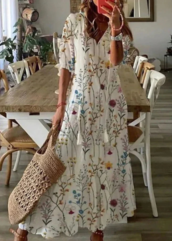 Frau in langem, geblümtem Kleid macht Selfie in modernem Esszimmer; Sommermode, Boho-Stil, Korbtasche, Holztisch, helle Einrichtung.