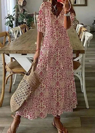 Frau in langem, gemustertem Sommerkleid mit Strohtasche steht in Esszimmer. Holztisch und Stühle im Hintergrund. Boho-Chic, Mode, Inneneinrichtung.