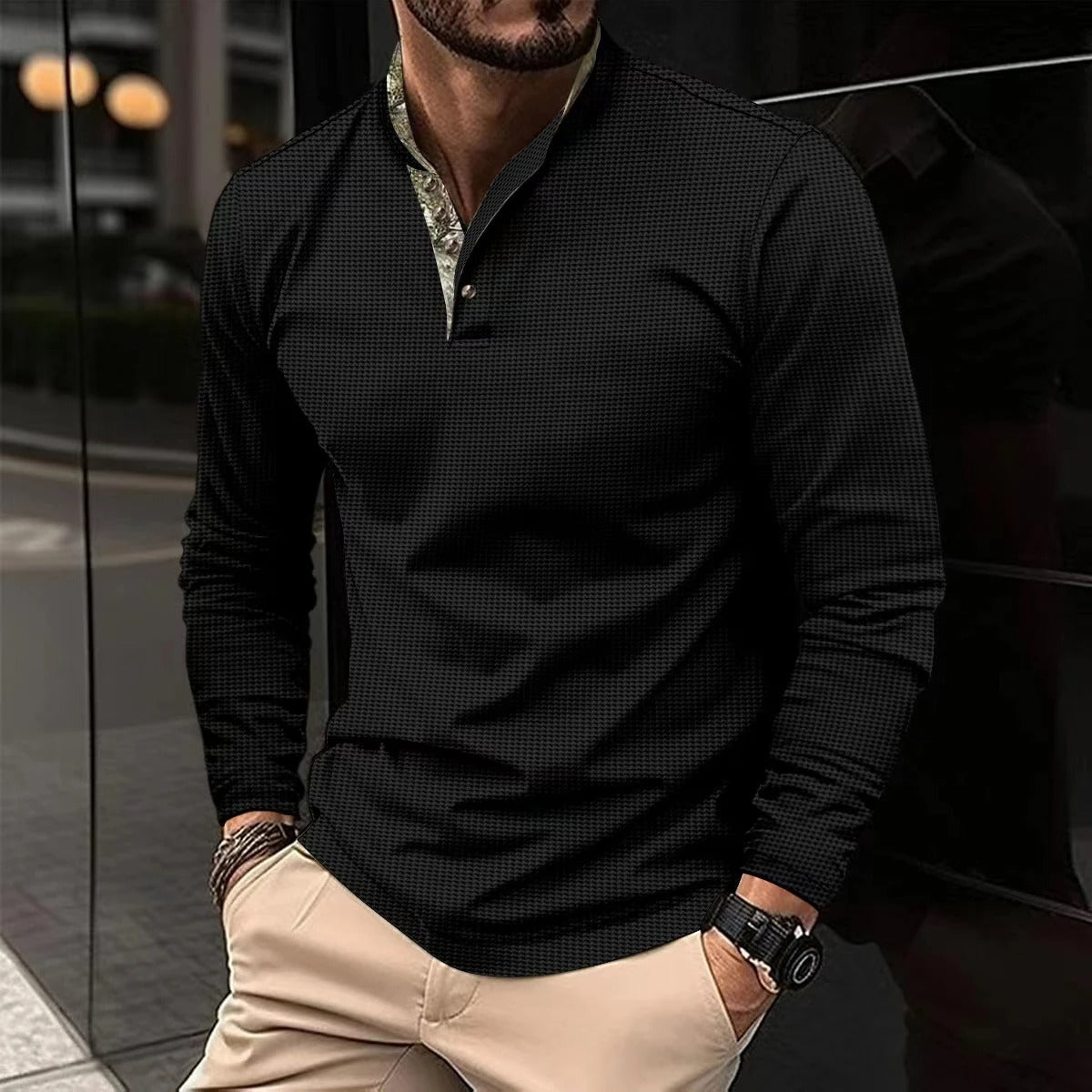 Mann in schwarzem Langarmhemd und beigen Hosen, steht vor einem modernen Gebäude. Stilvolle Herrenmode, elegantes Freizeitoutfit, urbane Kulisse.
