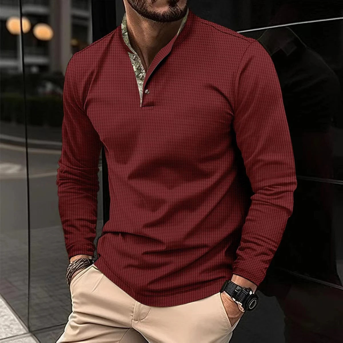 Mann in rotem Langarmhemd mit Stehkragen, kombiniert mit beigen Hosen, steht lässig an einer Wand. Elegante Herrenmode, urbaner Stil.