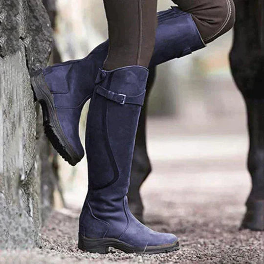 Braune Reitstiefel aus Leder an einer Person, die neben einem Pferd steht. Outdoor-Reitbekleidung, robustes Design, ideal für Reitsport.