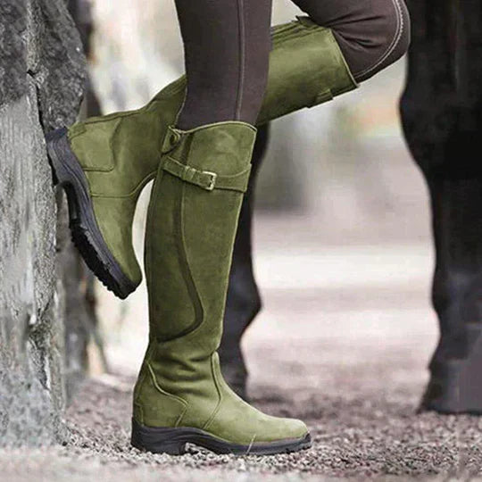 Grüne Reitstiefel aus Wildleder an einer Person, die an einer Steinmauer lehnt. Outdoor-Mode, langlebige Stiefel, stilvolle Reitbekleidung.
