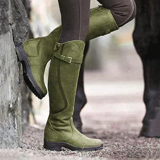 Grüne Reitstiefel aus Wildleder an einer Person, die an einer Steinmauer lehnt. Outdoor-Mode, langlebige Stiefel, stilvolle Reitbekleidung.