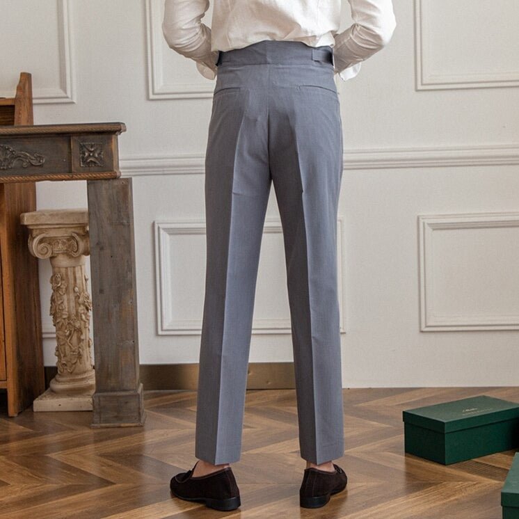 Mann in grauer Anzughose, weißem Hemd, steht in elegantem Raum mit Holzboden und antikem Tisch. Businessmode, Herrenbekleidung, stilvoll.