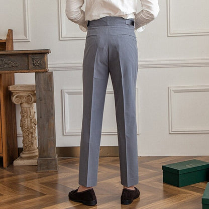 Mann in grauer Anzughose, weißem Hemd, steht in elegantem Raum mit Holzboden und antikem Tisch. Businessmode, Herrenbekleidung, stilvoll.